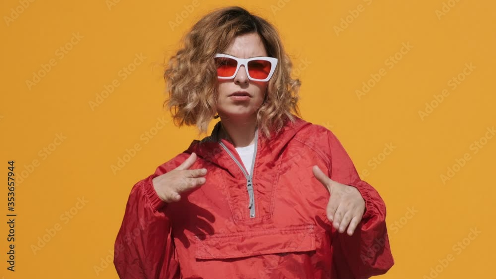 Portrait of happy woman in stylish sunglasses in red hoodie smiling, mimicking rap dance, rhythmically moving her hands to camera on yellow background slow motion. Emotions of people. Girl dancing Portrait of happy woman in stylish sunglasses in red hoodie smiling, mimicking rap dance, rhythmically moving her hands to camera on yellow background slow motion. Emotions of people. Girl dancing