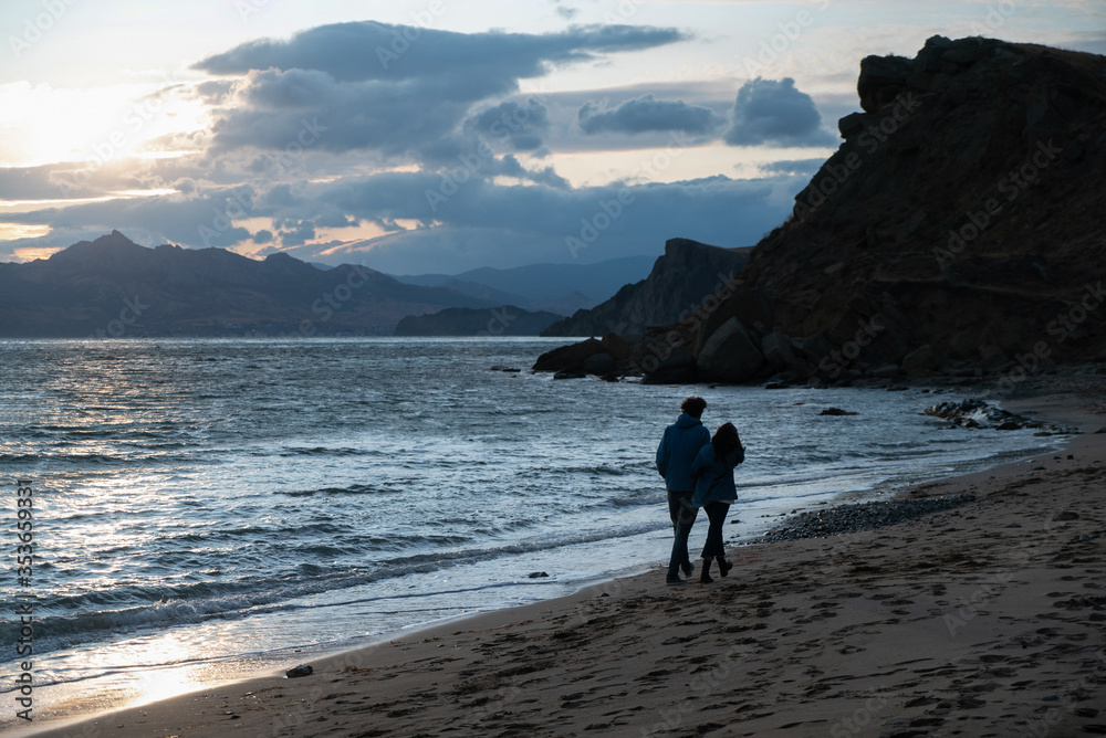 A couple of lovers walks hugging on the sandy beach along the rocky coast during sunset with views of clouds and mountains