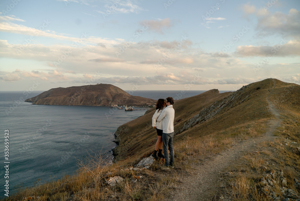 A couple of lovers warm each other with their arms, watching a panoramic view of the horizon and the coast under a beautiful blue sky with clouds