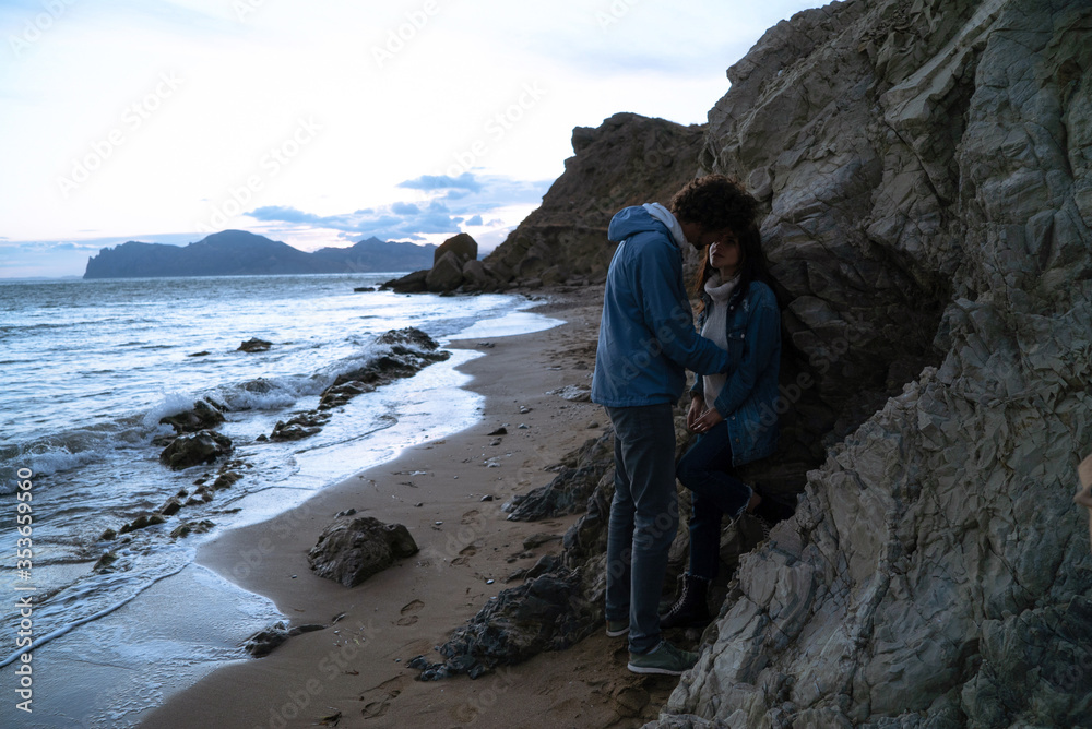 A couple of lovers hugs each other on the coast near the sea surf leaning against the beach rocks after sunset.