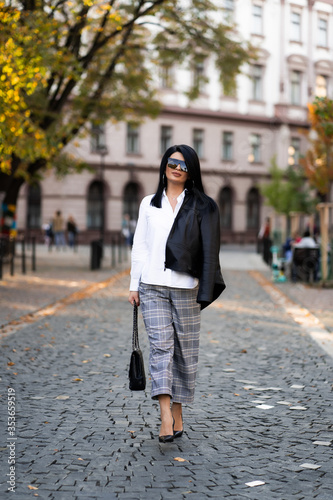 Autumn fashion outdoor. The girl in fashionable stylish white shirt , pants, jacket and sunglasses, autumnal lifestyle on the background of blurry yellow-green trees in the park. Vertical