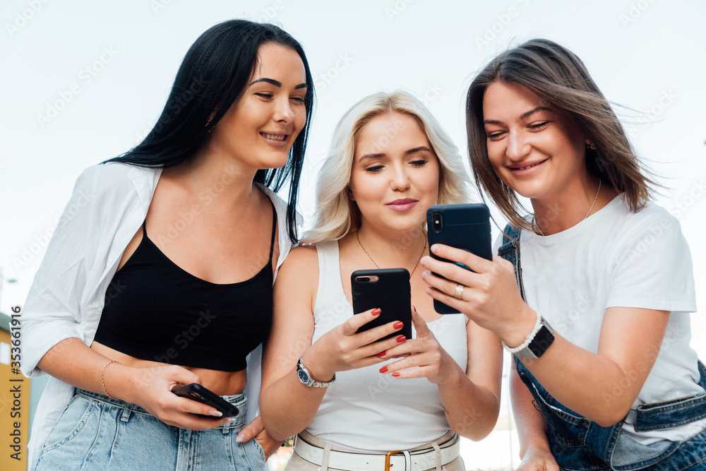 Group of three friends look at their smiley phones. The girls wear casual, color-neutral clothes. Group of three friends look at their smiley phones. The girls wear casual, color-neutral clothes.