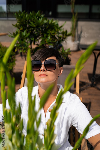 Beautiful woman with short brunette hair in white clothes and sunglasses. Fashion photography. Fashion model posing with flowers on wood background.