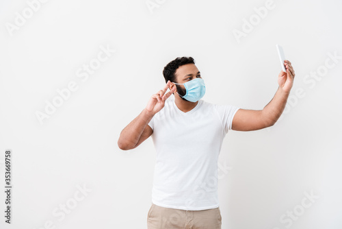 african american man in medical mask showing peace sign and taking selfie near white wall