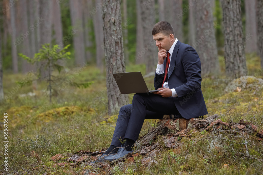 freelancing in nature. a man sits on a stump and works behind a laptop in the woods.