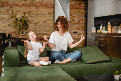 curly mother looking at daughter sitting in lotus pose on sofa