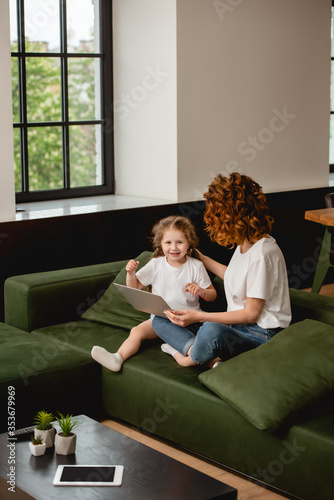 curly mother holding laptop near happy daughter in living room