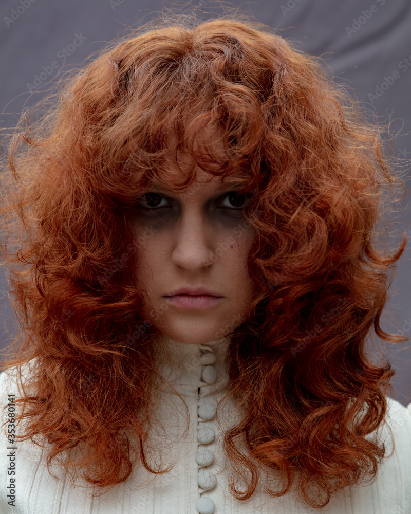 Fototapeta premium A portrait of a young woman with long curly, red hair, wearing a vintage dress and looking sad and serious.