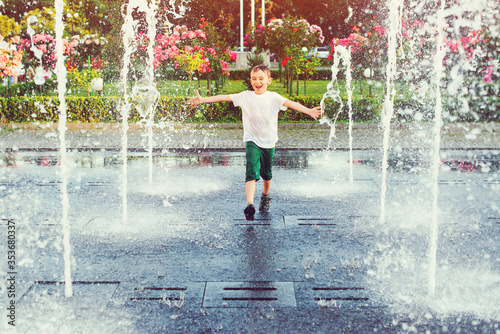 Happy boy running between water jets in city fountain. Summer vacation.