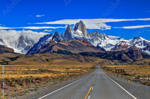 Fitz Roy and Torre mountains with route and cars at foreground, during a sunny day with clouds.