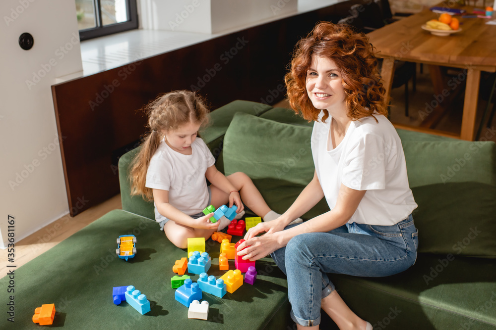 Obraz premium curly mother smiling near daughter playing building blocks in living room