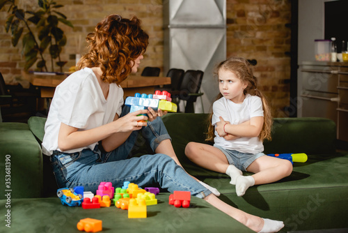 Foto curly mother holding toy gun and looking at offended daughter with crossed arms