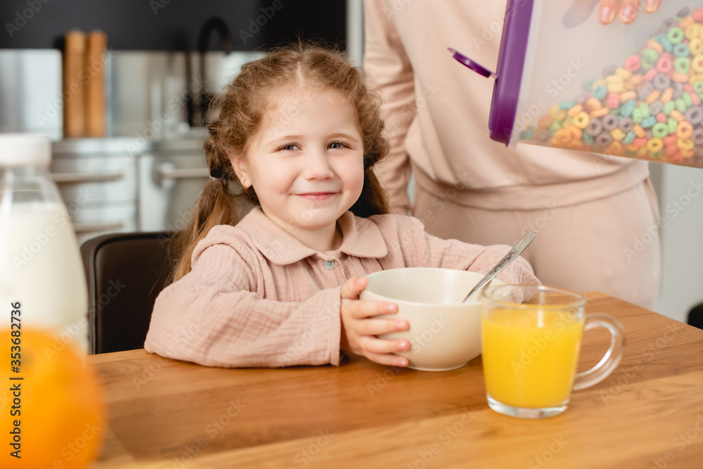 selective focus of mother holding container with delicious corn flakes near bowl and cute daughter selective focus of mother holding container with delicious corn flakes near bowl and cute daughter