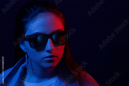 The young girl in a denim jacket and sunglasses bright neon blue and red light. The concept of beauty and fashion.
