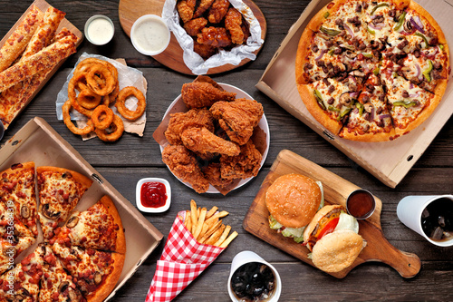 Buffet table scene of take out or delivery foods. Pizza, hamburgers, fried chicken and sides. Above view on a dark wood background. photo