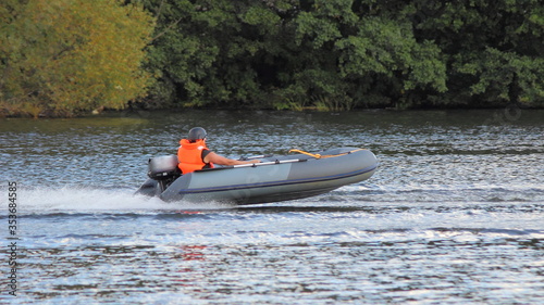 Grey Inflatable outboard motor boat with racer in brighy orange lifejacket and helmet fast glide floats on water Race at river on shore trees background at Sunny summer day, outdoor sport competition