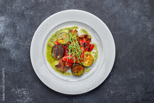 Grilled baked vegetables, eggplant, zucchini, bell pepper, champignon mushrooms, on a white plate against a dark background, with microgreen