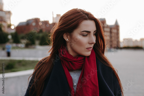 Beautiful young woman in black wool coat and in red scarf looking sideways with sad look and thinking.
Stylish girl on the city background