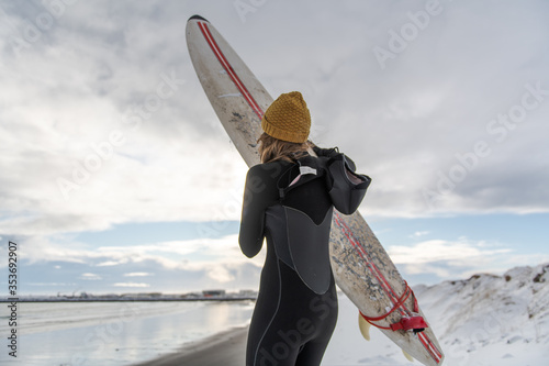 Rear view of a woman wearing a wetsuit and holding a surfboard standing on a snowy beach and looking out to sea.