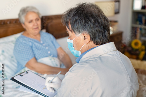 A doctor in a white coat and protective face mask making a home visit to a senior woman patient. photo