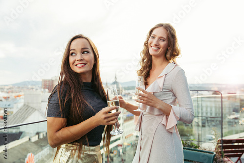 Two women dressed to go out for the evening standing on a balcony overlooking a city holding glasses of champagne.
