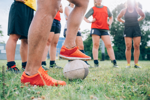 The legs and feet of a group of women standing on a training pitch one with a foot on a rugby ball. photo