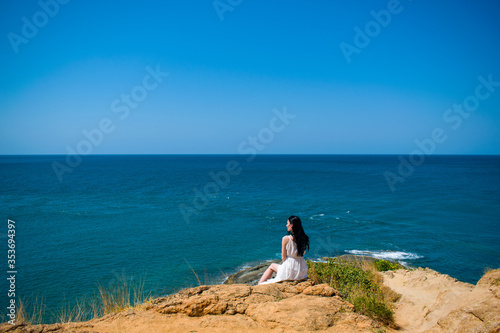 A romantic young girl brunette in a long white dress sits on the edge of the island  s land. Beautiful landscape of exotic white sand sea beach Southeast Asia  Thailand. View from the mountain.