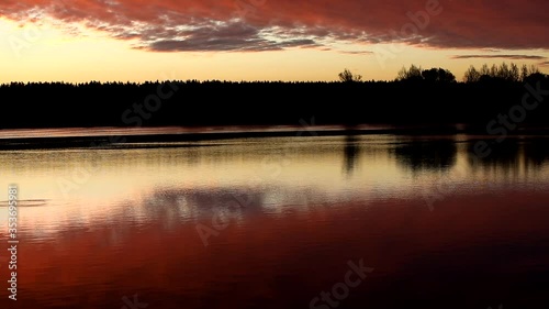 Mirror image of pink light sky with bright burning crimson clouds in the lake at dawn.The illuminated black forest on the horizon is copied in the calm water.A glossy picture in the twilight.Russia