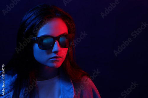 The young girl in a denim jacket and sunglasses bright neon blue and red light. The concept of beauty and fashion.