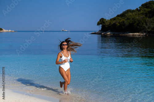 Young girl running on beach. Athletic happy woman jogging in trendy sexy white bodysuit enjoying the sun exercising. Healthy lifestyle. Fun walk along the shore. Perfect fitness body shapes and tan