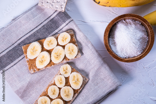 Healthy snack with Swedish rye crispbread, peanut butter and banana's slices on a cloth and a wooden bowl with grated coconut. Top view. Flat lay.