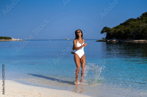 Fototapeta Naklejka Na Ścianę i Meble -  Young girl running on beach. Athletic happy woman jogging in trendy sexy white bodysuit enjoying the sun exercising. Healthy lifestyle. Fun walk along the shore. Perfect fitness body shapes and tan