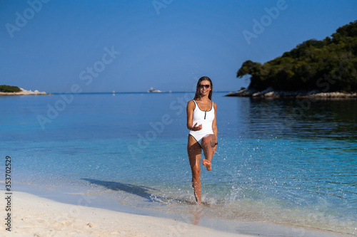 Young girl running on beach. Athletic happy woman jogging in trendy sexy white bodysuit enjoying the sun exercising. Healthy lifestyle. Fun walk along the shore. Perfect fitness body shapes and tan