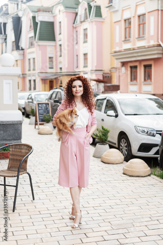 Close up portrait of young beautiful fashionable redhead young woman, long curly hair, wearing pink suit, posing in street of european city.