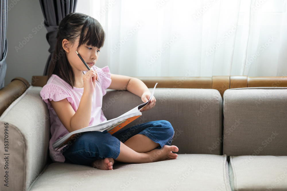 Asian little girl thinking while reading a book on the sofa in the room. Asian little girl thinking while reading a book on the sofa in the room.