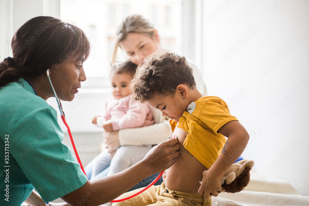 Side view of female doctor examining boy's heartbeat with stethoscope while mother sitting and daughter in clinic Side view of female doctor examining boy's heartbeat with stethoscope while mother sitting and daughter in clinic