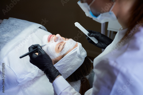 A female cosmetologist side view who applies peeling gommage on the girl’s face . photo