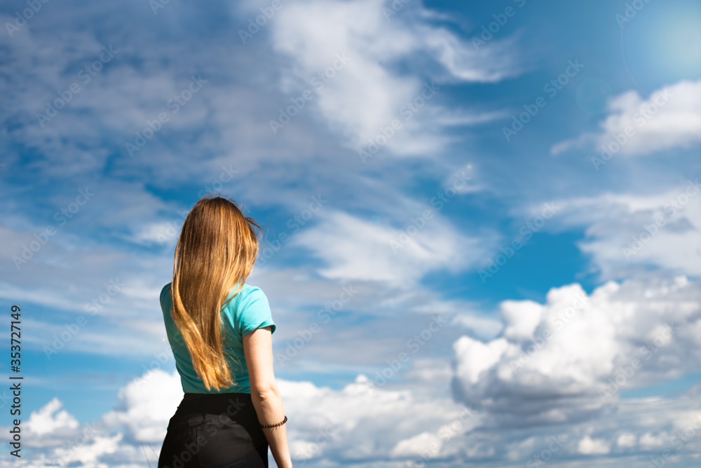 woman with blond hair stands with her back and looks into the distance, against the background of blue sky and white clouds woman with blond hair stands with her back and looks into the distance, against the background of blue sky and white clouds