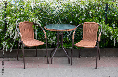 Summer street cafe. The glass table and brown chairs are outdoors. A small cozy restaurant with retro furniture and bushes with white flowers.