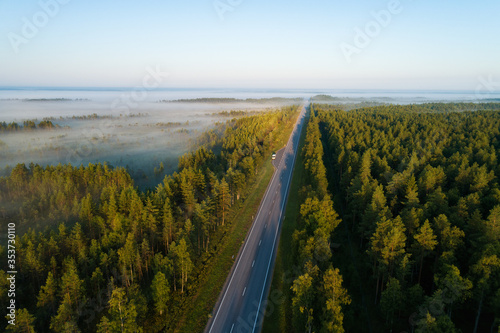 Aerial top view of highway road. Summer forest. Early morning, sunrays, foggy road. Motorway trough the countryside at the sunrise.