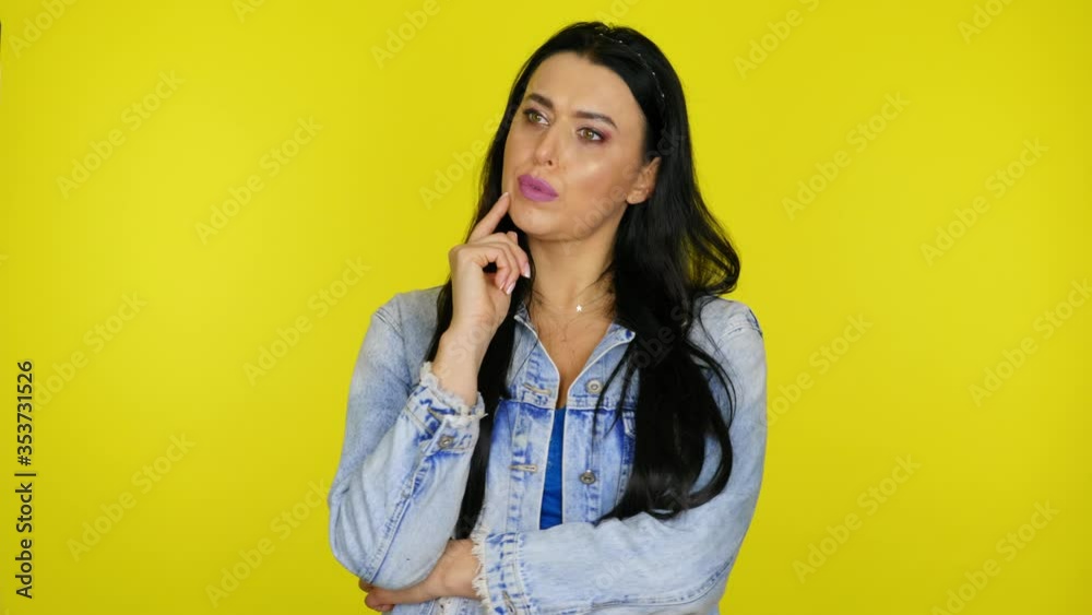 Serious woman with black hair in a denim jacket looks around and thinks what to choose by leaning her finger on her chin on a yellow background with copy space. Place for text or product. 4K footage