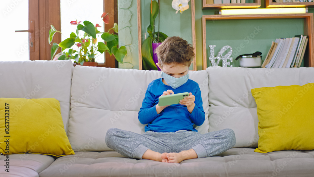 At home in quarantine concentrated small boy with a protective mask playing the games on the smartphone while sitting on the sofa Coronavirus 2019 At home in quarantine concentrated small boy with a protective mask playing the games on the smartphone while sitting on the sofa Coronavirus 2019