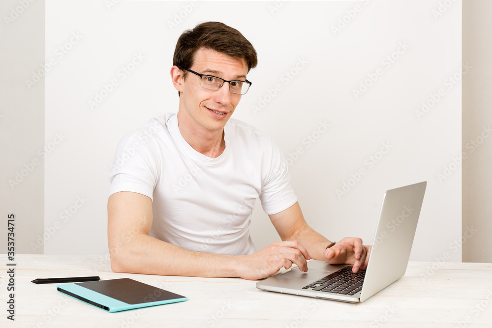 A handsome man with a light smile wearing a T-shirt and glasses sits against a white wall at his desk and work with a laptop. The concept of remote quarantine education and communication A handsome man with a light smile wearing a T-shirt and glasses sits against a white wall at his desk and work with a laptop. The concept of remote quarantine education and communication