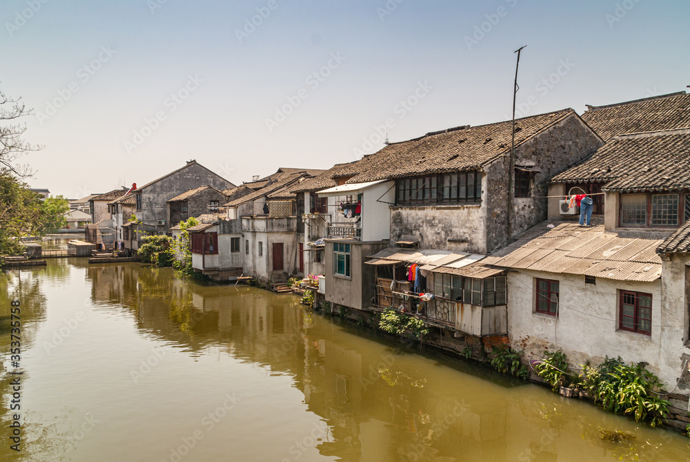 Tongli, JIangsu, China - May 3, 2010: Line of dark roofed dirty poor white houses reflected in greenish water of canal under light blue sky. Green foliage and laundry add color.