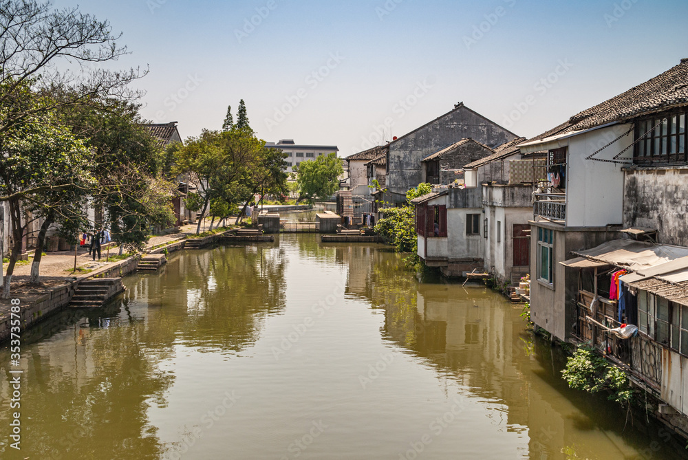 Tongli, JIangsu, China - May 3, 2010: 2 Lines of dark roofed dirty poor white houses reflected in greenish water of canal under light blue sky. Green foliage and laundry add color. Tongli, JIangsu, China - May 3, 2010: 2 Lines of dark roofed dirty poor white houses reflected in greenish water of canal under light blue sky. Green foliage and laundry add color.