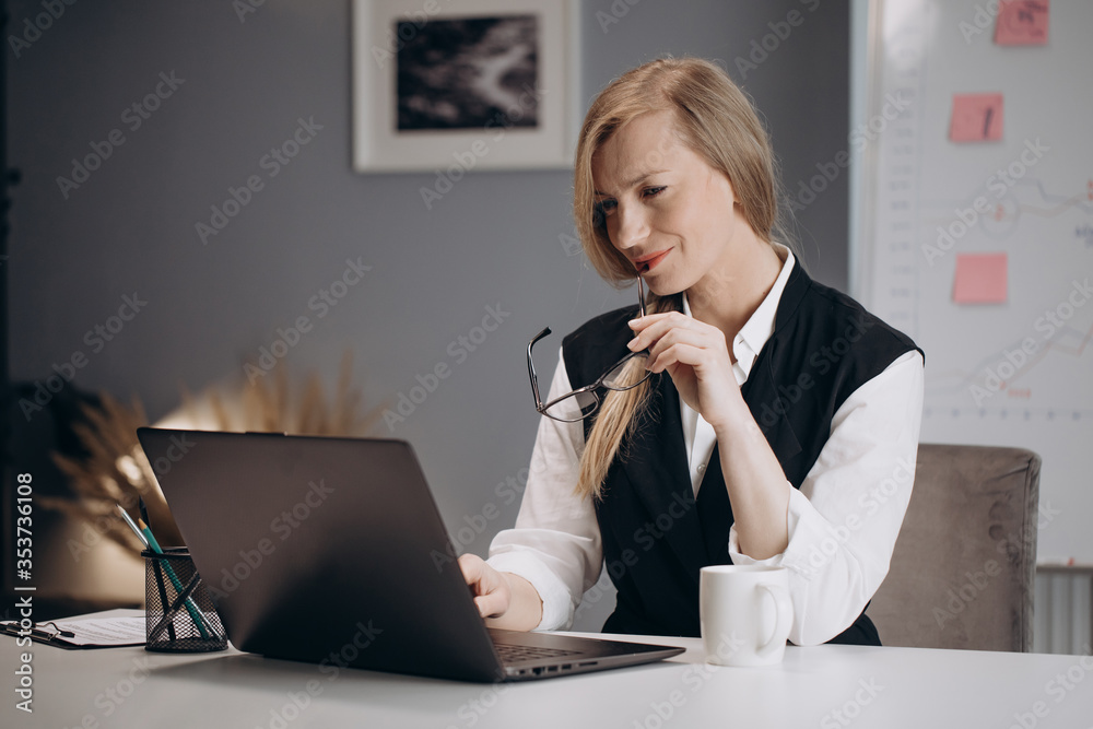 Smart business woman with blond hair sitting at table and looking at computer screen. Mature lady in formal outfit working at modern office center Smart business woman with blond hair sitting at table and looking at computer screen. Mature lady in formal outfit working at modern office center