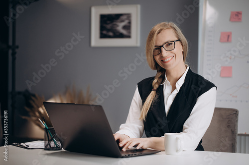 Charming woman in eyeglasses and stylish outfit sitting at table with opened laptop, smiling and looking at camera. Concept of working process and business career