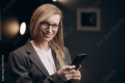 Charming businesswoman wearing glasses and formal suit holding smartphone while looking at camera. Happy lady with blond hair relaxing after work at own office.