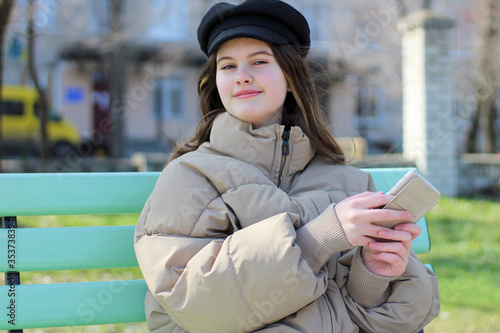 Satisfied smiling girl in a black cap and with a phone in her hands sits on a bench and looks at the camera