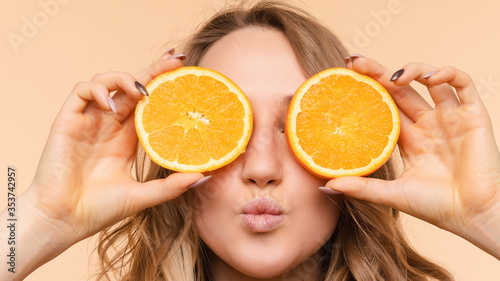 Front view of cheerful young blonde posing with fresh oranges on isolated background. Funny girl keeping fruit and closing eyes in studio. Concept of happiness and health.